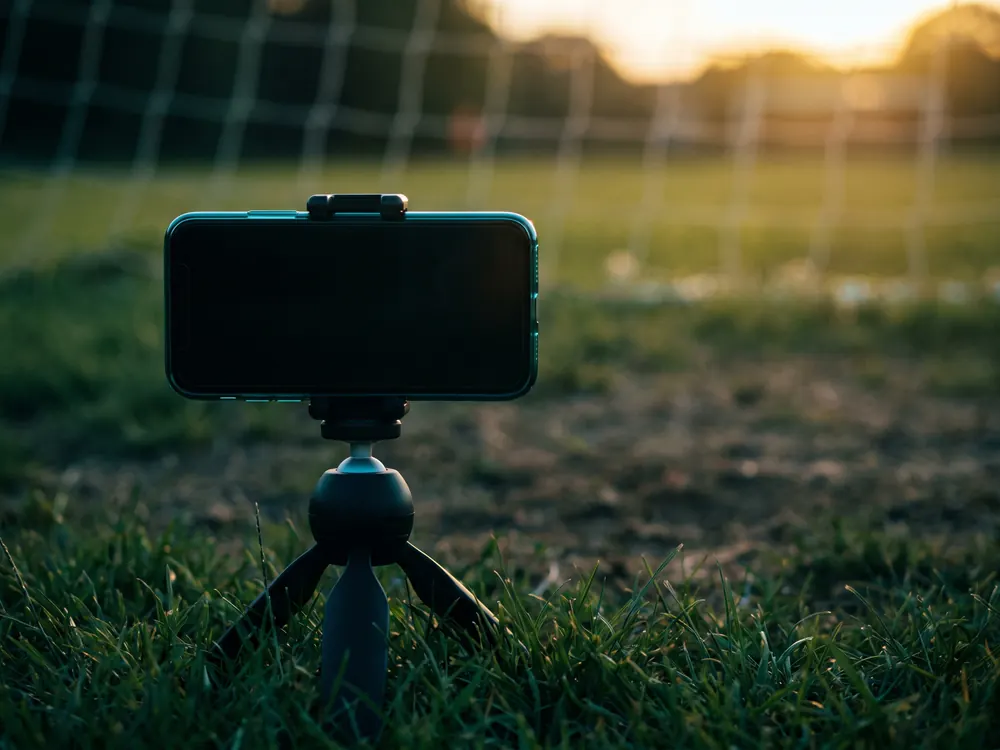 Un celular acostado sobre un trípode chico, apoyado en el pasto al borde de la cancha.